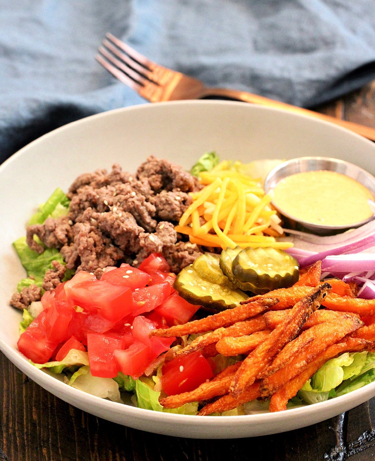 A burger bowl on a wooden board with a blue napkin and bronze fork nearby.