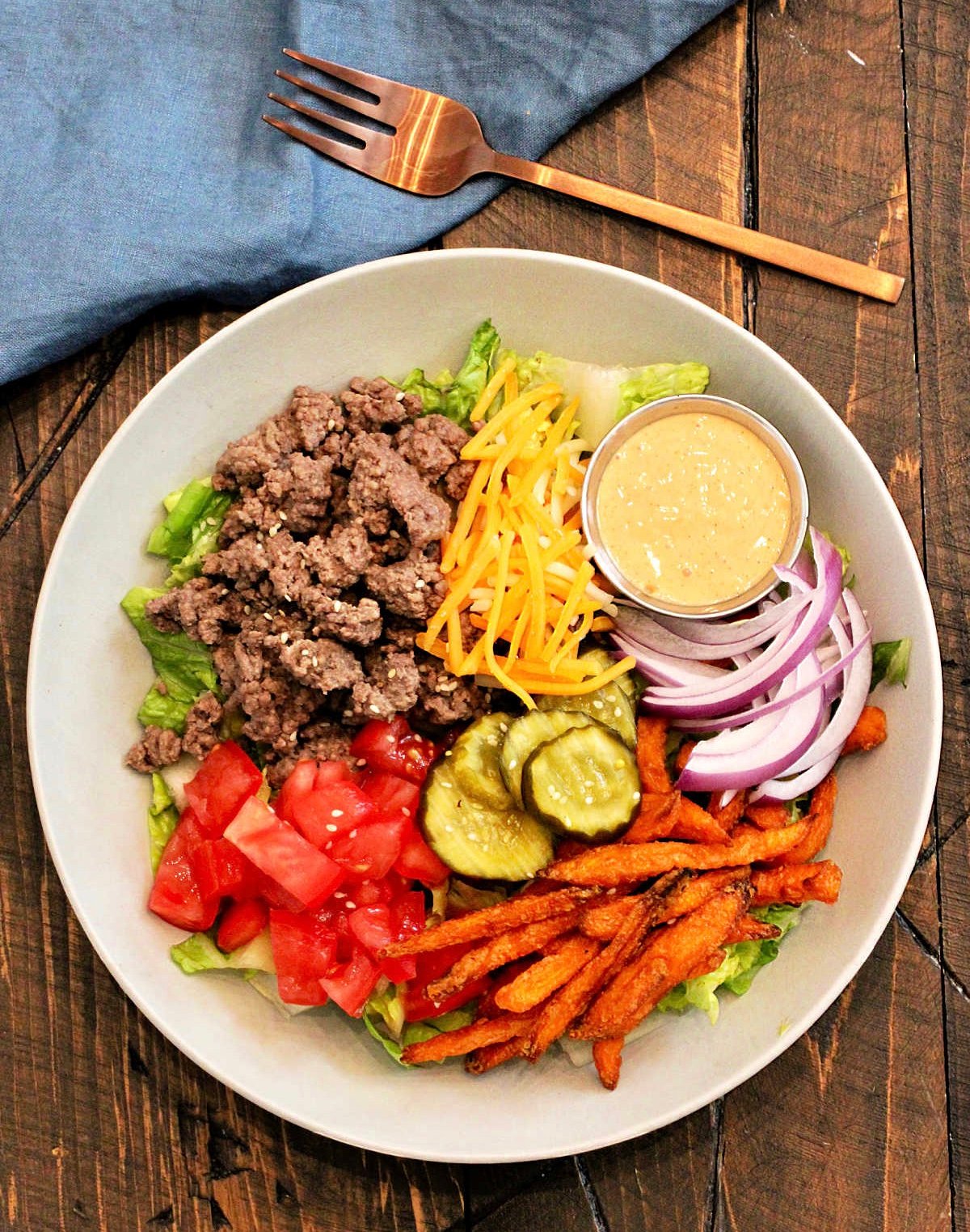 A burger bowl on a wooden board with a blue napkin and bronze fork nearby.