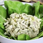Pesto chicken salad on a bed of lettuce in a white bowl with a gold spoon nearby.