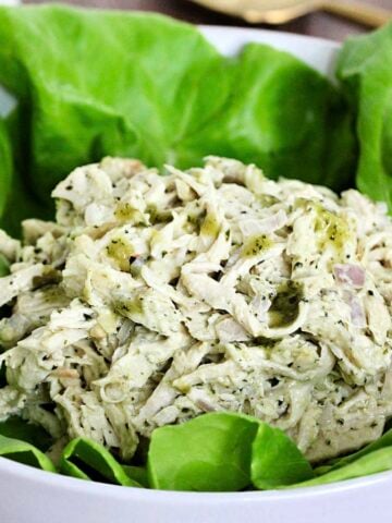 Pesto chicken salad on a bed of lettuce in a white bowl with a gold spoon nearby.