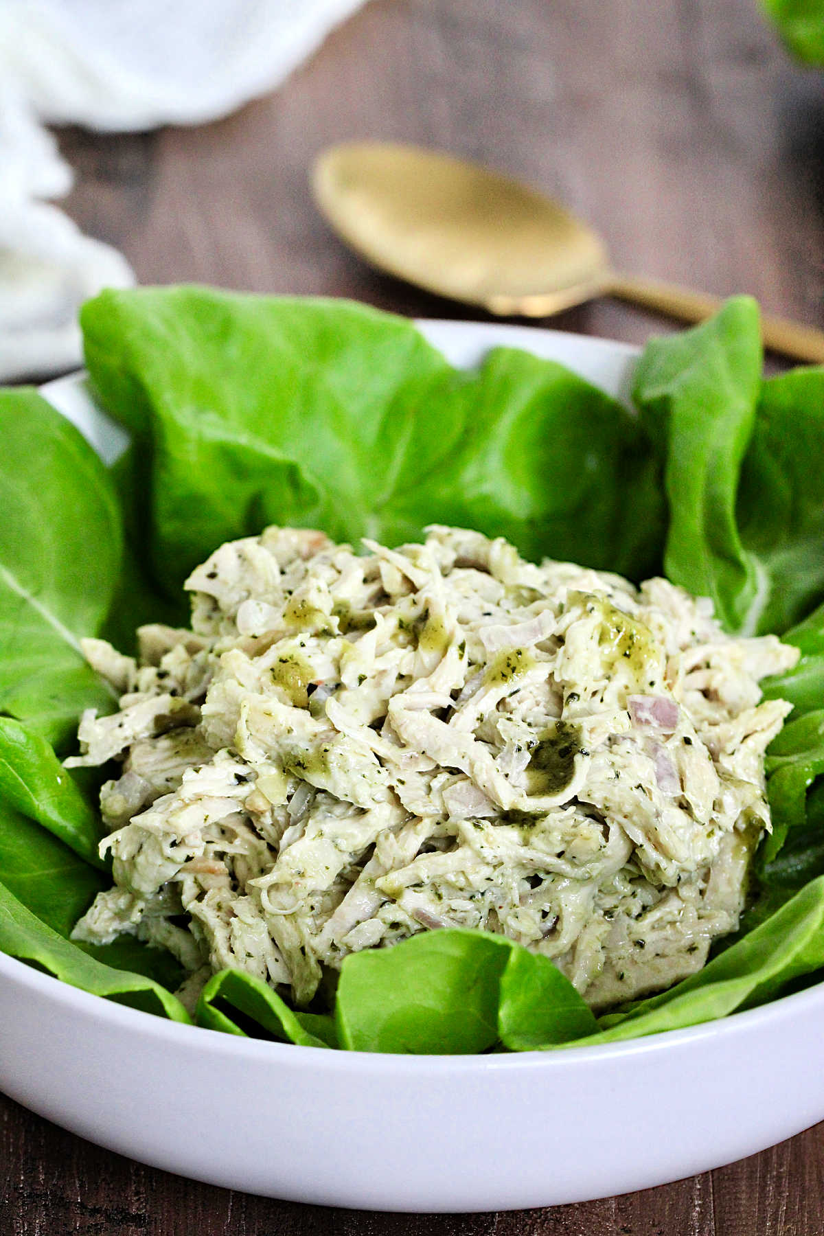 Pesto chicken salad on a bed of lettuce in a white bowl with a gold spoon nearby.