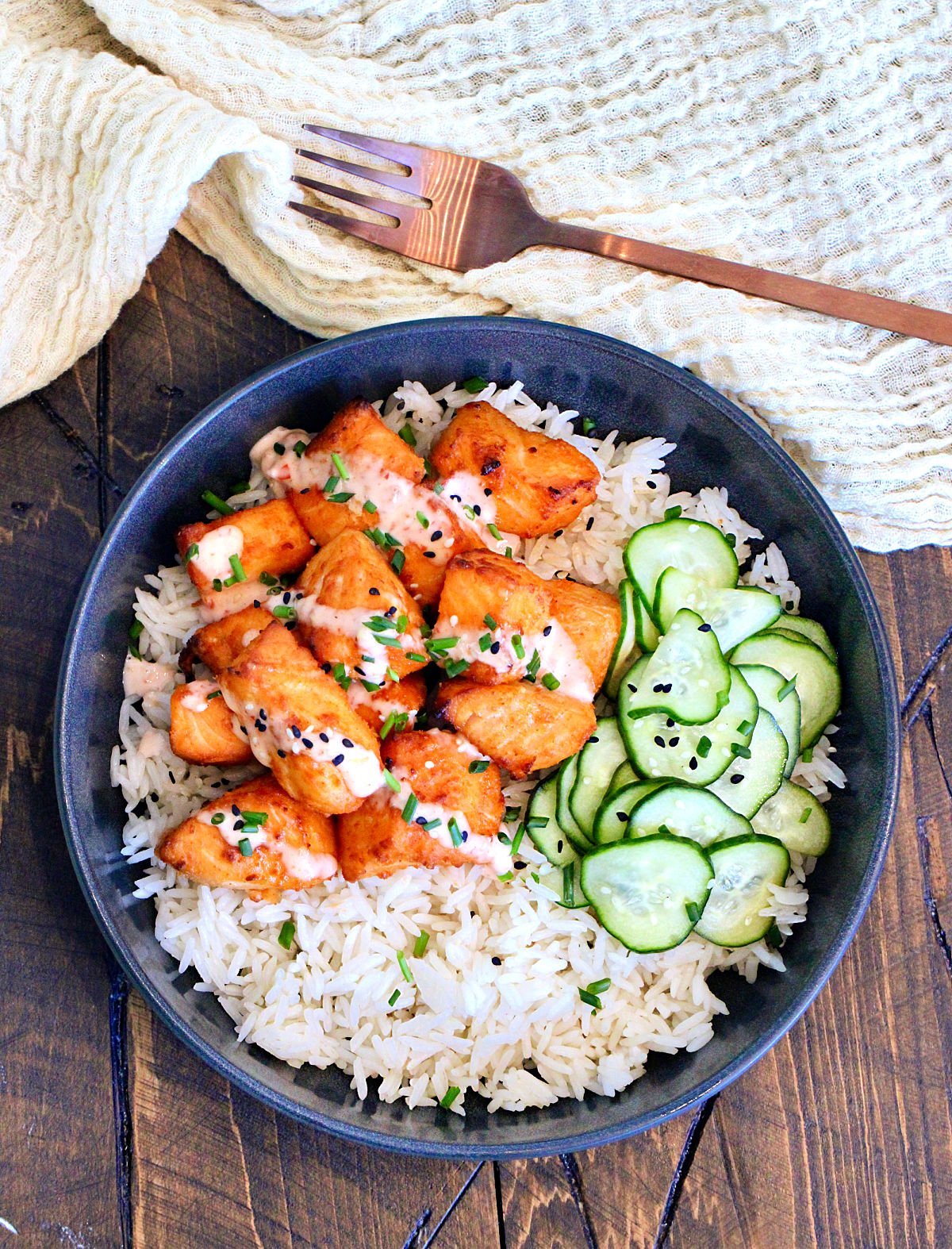 Bang Bang Salmon Bites with pickled cucumbers on a bed of white rice in a black bowl.