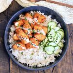 Bang Bang Salmon Bites with pickled cucumbers on a bed of white rice in a black bowl.