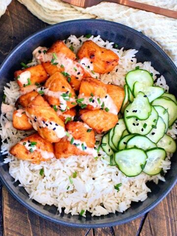 Bang Bang Salmon Bites with pickled cucumbers on a bed of white rice in a black bowl.