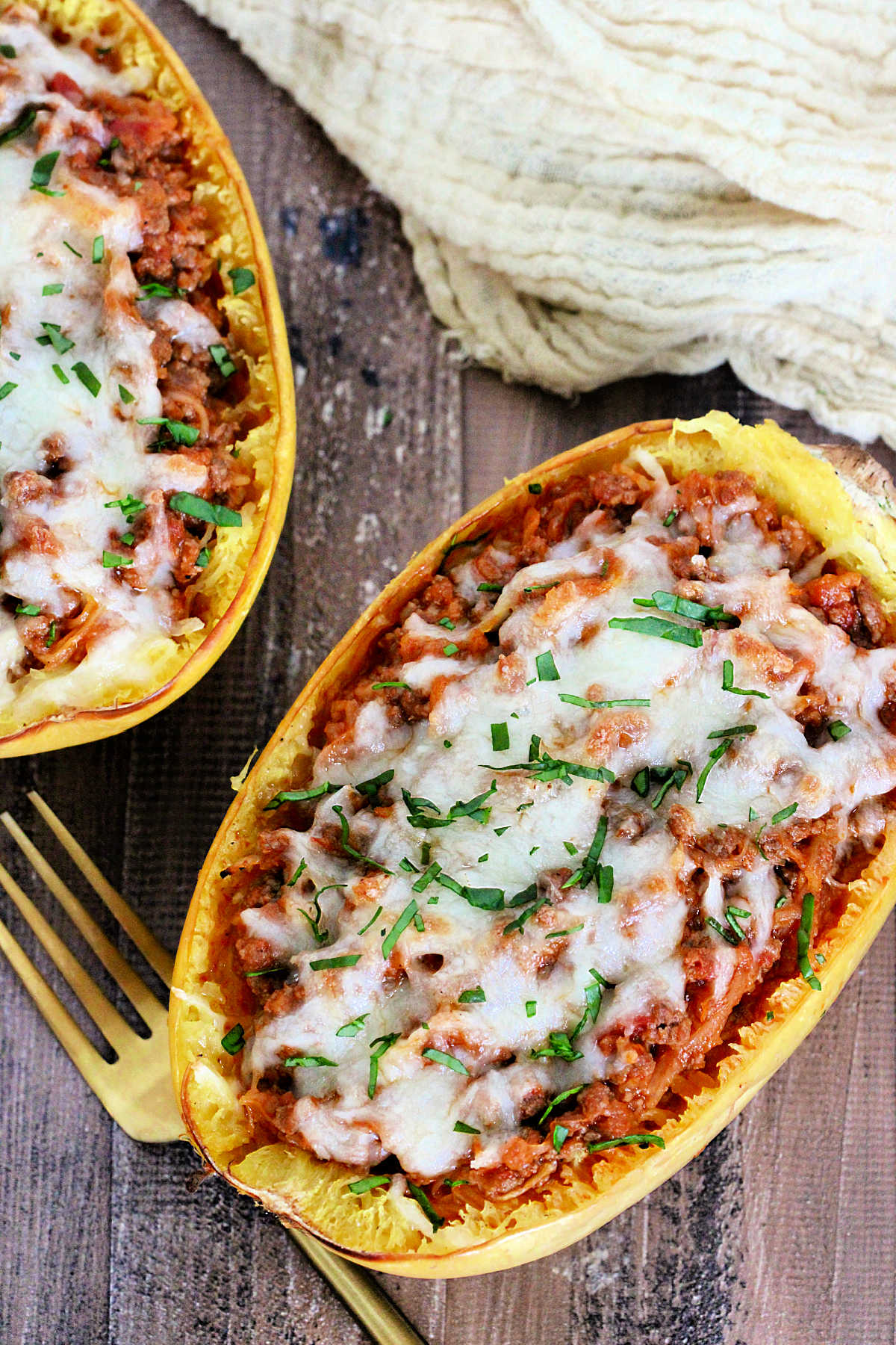 Overhead of 2 spaghetti squash boats on a wooden board with a tan napkin and gold fork nearby.