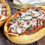 2 spaghetti squash boats on a wooden board with a tan napkin and gold fork nearby.