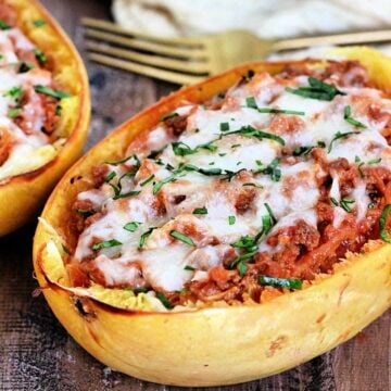 2 spaghetti squash boats on a wooden board with a tan napkin and gold fork nearby.
