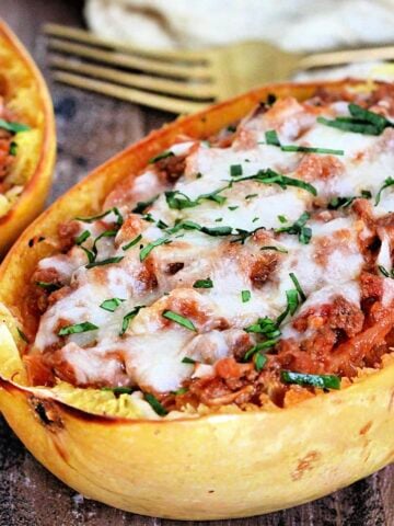 2 spaghetti squash boats on a wooden board with a tan napkin and gold fork nearby.