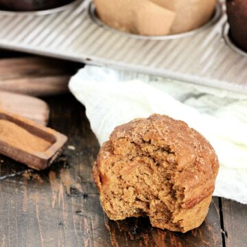 Easy Pumpkin Banana Muffin with a bite out of it near a muffin pan and wooden measuring spoons.
