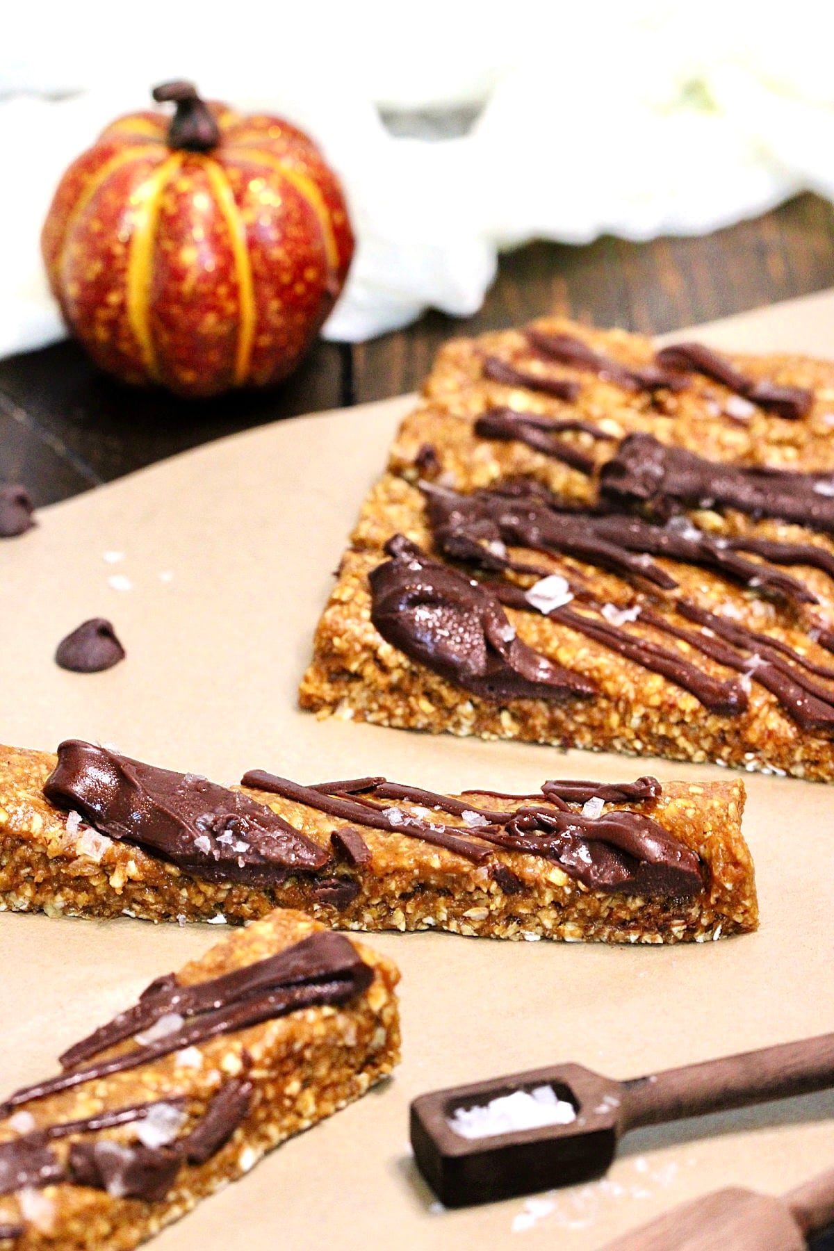 Pumpkin protein bars on parchment paper surrounded by chocolate chips, measuring spoons and a small pumpkin.