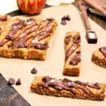 Pumpkin protein bars on parchment paper surrounded by chocolate chips, measuring spoons and a small pumpkin.