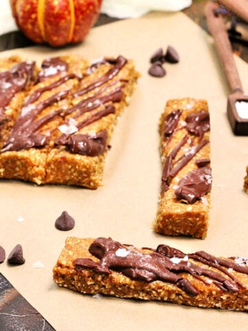 Pumpkin protein bars on parchment paper surrounded by chocolate chips, measuring spoons and a small pumpkin.