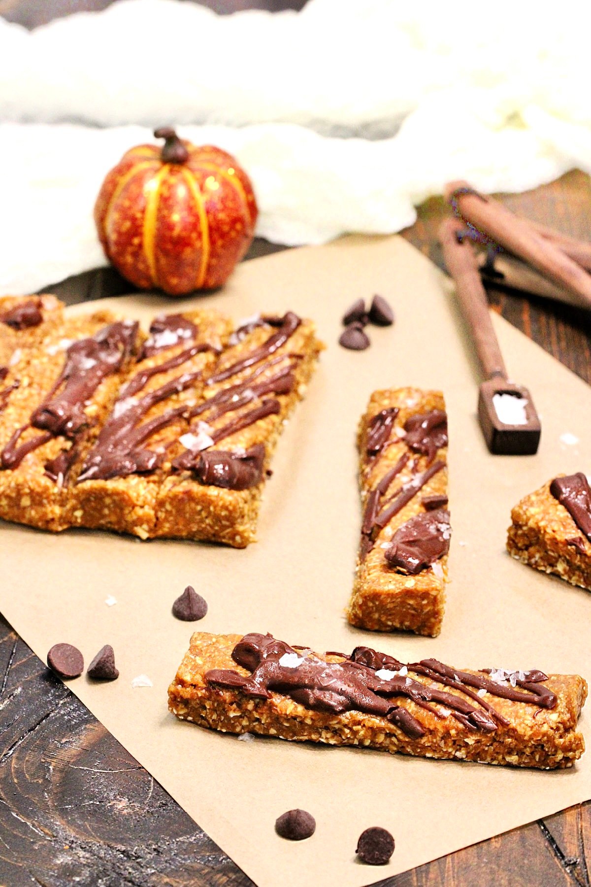 Pumpkin protein bars on parchment paper surrounded by chocolate chips, measuring spoons and a small pumpkin.