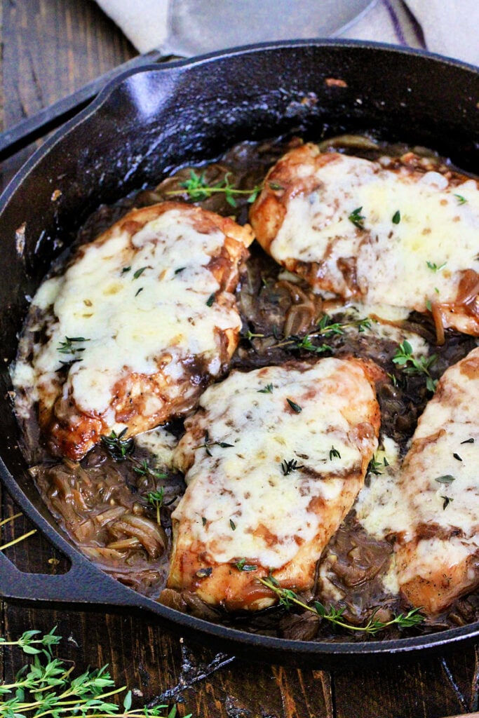 Easy Skillet French Onion Chicken in a cast iron skillet on a wooden board surround by fresh thyme and a purple napkin.