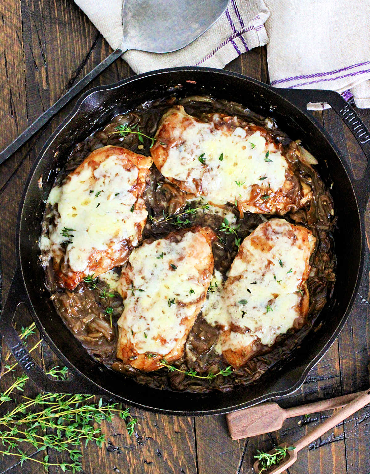 Easy Skillet French Onion Chicken in a cast iron skillet on a wooden board surround by fresh thyme, wooden measuring spoons and a purple napkin.