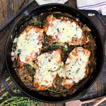 Easy Skillet French Onion Chicken in a cast iron skillet on a wooden board surround by fresh thyme, wooden measuring spoons and a purple napkin.