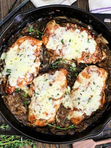 Easy Skillet French Onion Chicken in a cast iron skillet on a wooden board surround by fresh thyme, wooden measuring spoons and a purple napkin.