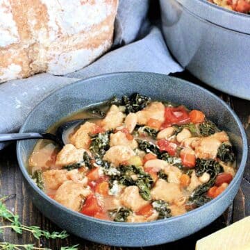 A bowl of Tuscan ribollita on a wooden board surrounded by a loaf of bread, thyme, Parmesan and a dutch oven.