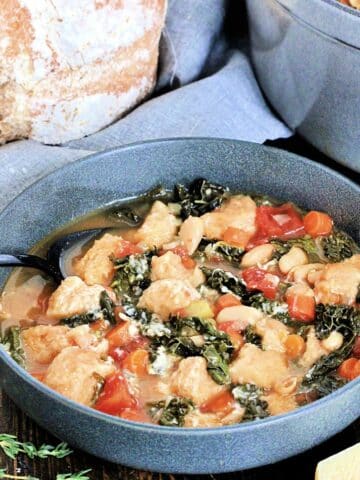 A bowl of Tuscan ribollita on a wooden board surrounded by a loaf of bread, thyme, Parmesan and a dutch oven.