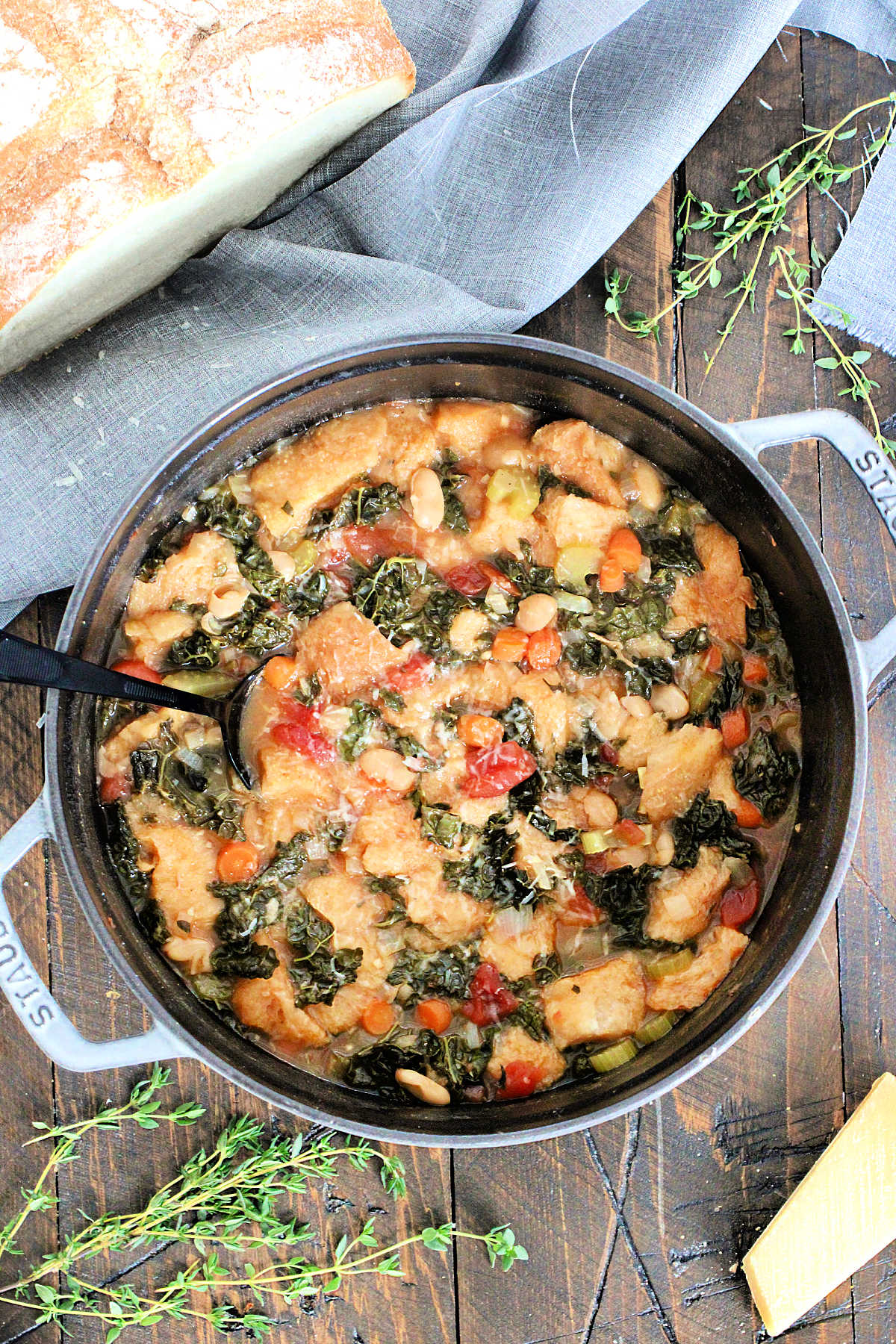 A Dutch oven full of Tuscan ribollita on a wooden board surrounded by a loaf of bread, thyme, and a chunk of Parmesan cheese.