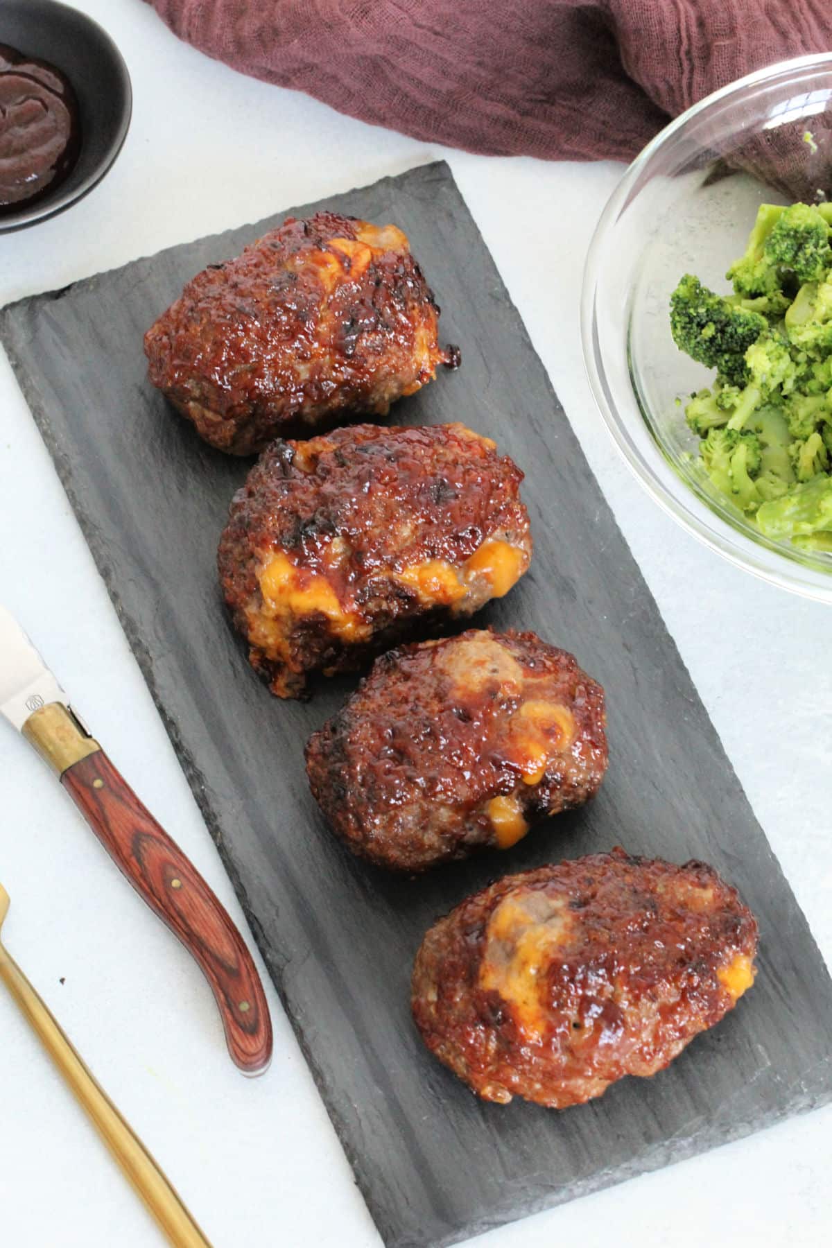 Overhead of barbecue cheddar mini meatloaves on slate board with bowl of broccoli nearby.
