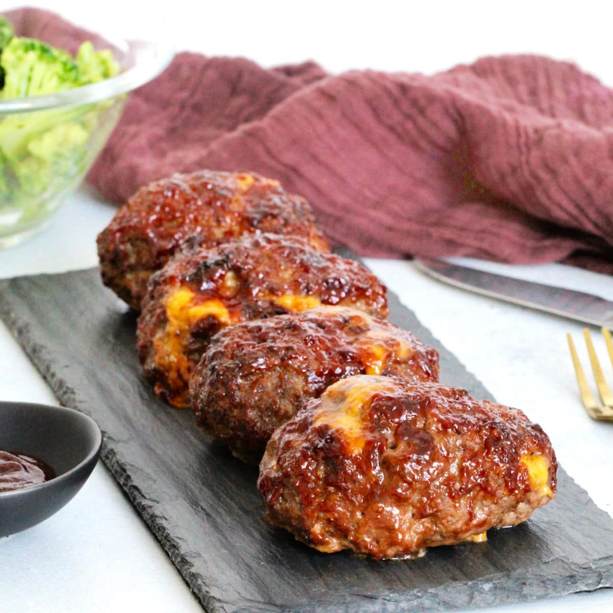 Side view of barbecue cheddar mini meatloaves on slate board with bowl of broccoli nearby.