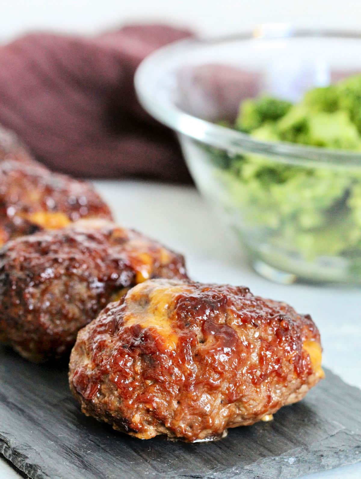 Side view of barbecue cheddar mini meatloaves on slate board with bowl of broccoli nearby.