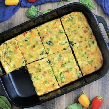 Overhead view of high protein cottage cheese egg bake in a black rectangular baking dish surrounded by a blue napkin, colorful peppers and spinach leaves.