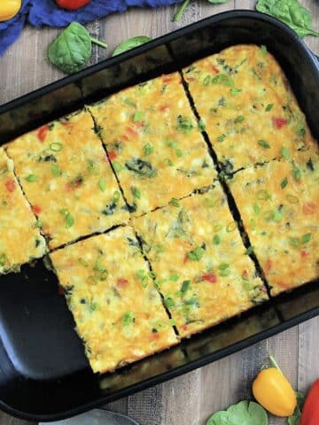 Overhead view of high protein cottage cheese egg bake in a black rectangular baking dish surrounded by a blue napkin, colorful peppers and spinach leaves.
