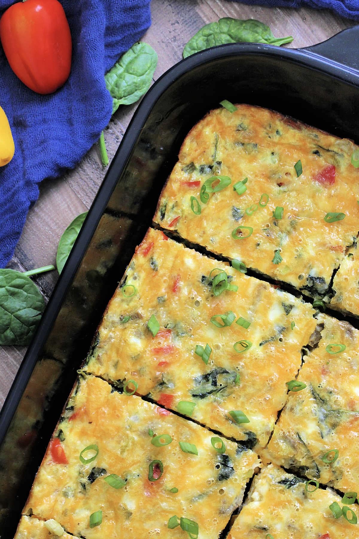 Overhead view of high-protein egg casserole with cottage cheese in a black baking dish surrounded by a blue napkin, colorful peppers and spinach leaves.