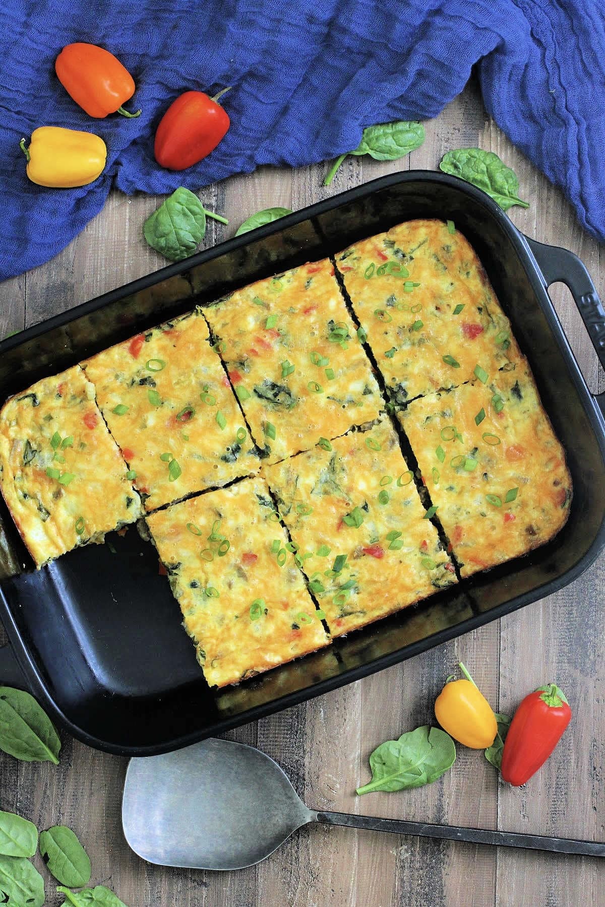 Overhead view of high protein cottage cheese egg bake in a black rectangular baking dish surrounded by a blue napkin, colorful peppers and spinach leaves.