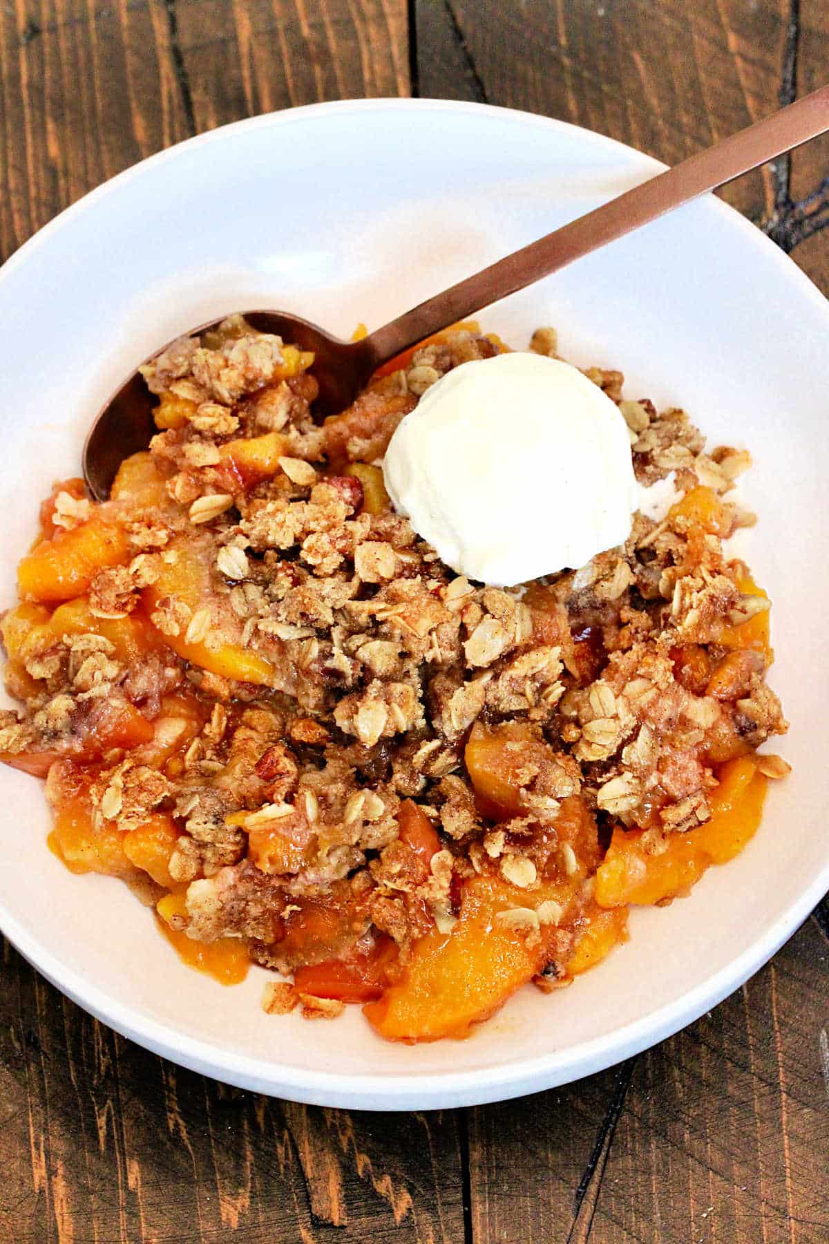 Gluten free peach crisp in a white bowl on a wooden board with a scoop of vanilla ice cream in the bowl.