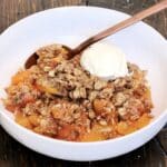 Gluten free peach crisp in a white bowl on a wooden board with a scoop of vanilla ice cream in the bowl.