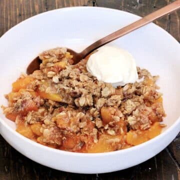 Gluten free peach crisp in a white bowl on a wooden board with a scoop of vanilla ice cream in the bowl.